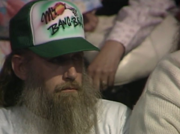 A bearded manwearing a sick airbrushed hat that reads "Mr. Bang Bang" with a firing revolver on it.